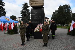 Uczniowie w mundurach, stojący przed pomnikiem, obok biało-czerwone flagi