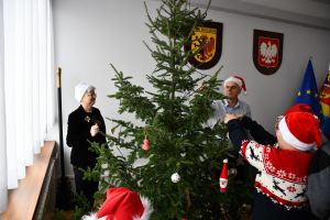 Dzieci z Panią sekretarz i starostą, w czapkach Mikołaja ubierają choinkę