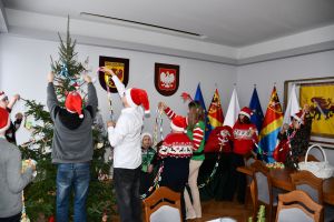 Dzieci z Panią sekretarz i starostą, w czapkach Mikołaja ubierają choinkę