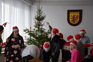Dzieci z Panią sekretarz i starostą, w czapkach Mikołaja ubierają choinkę